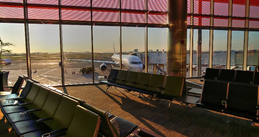 The sun rises over an empty gate at Boston Logan International Airport, a passenger plane visible outside the floor-to-ceiling windows